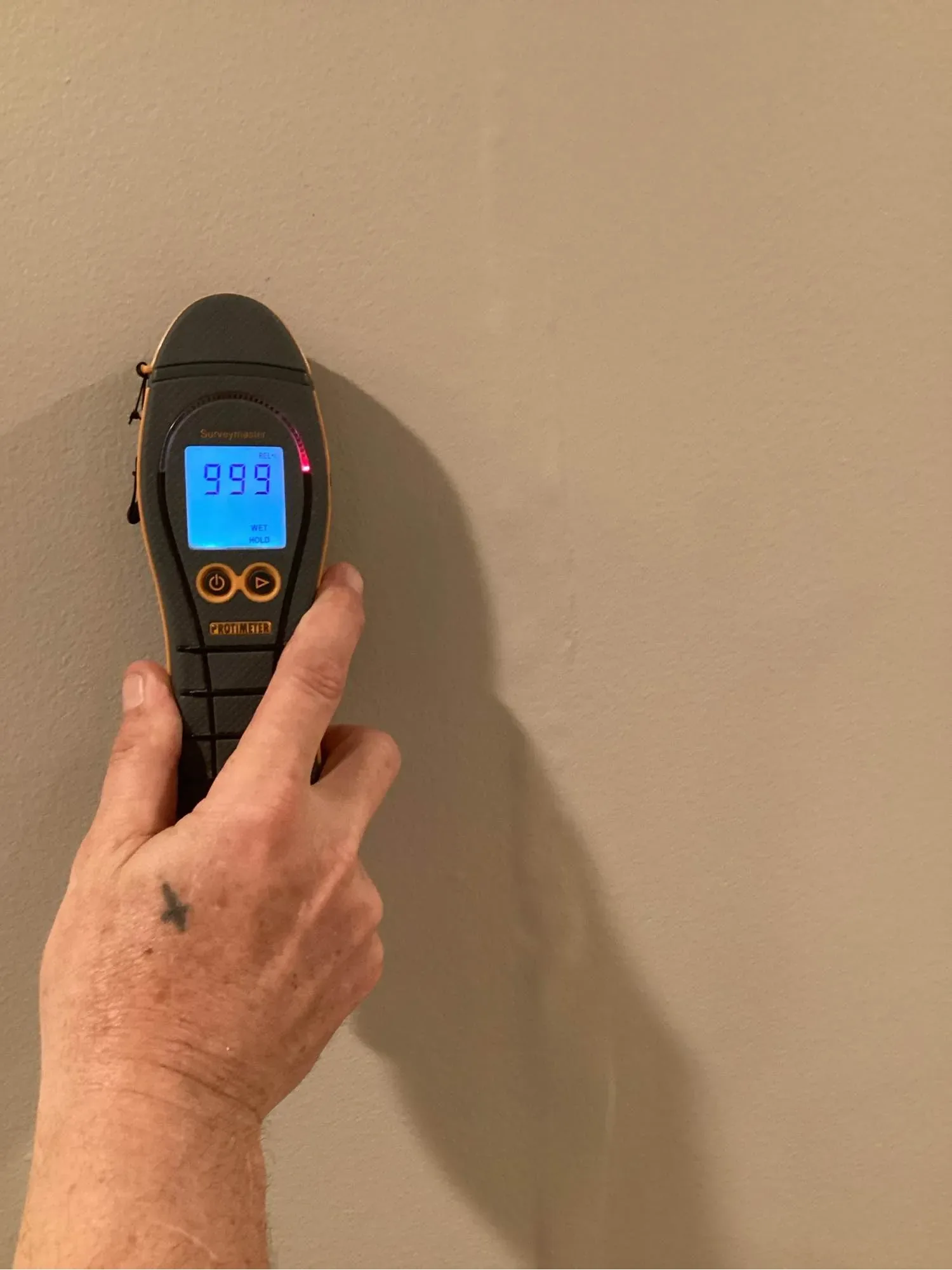 A hand holding a moisture meter flat against a wall, with the reading showing on the display