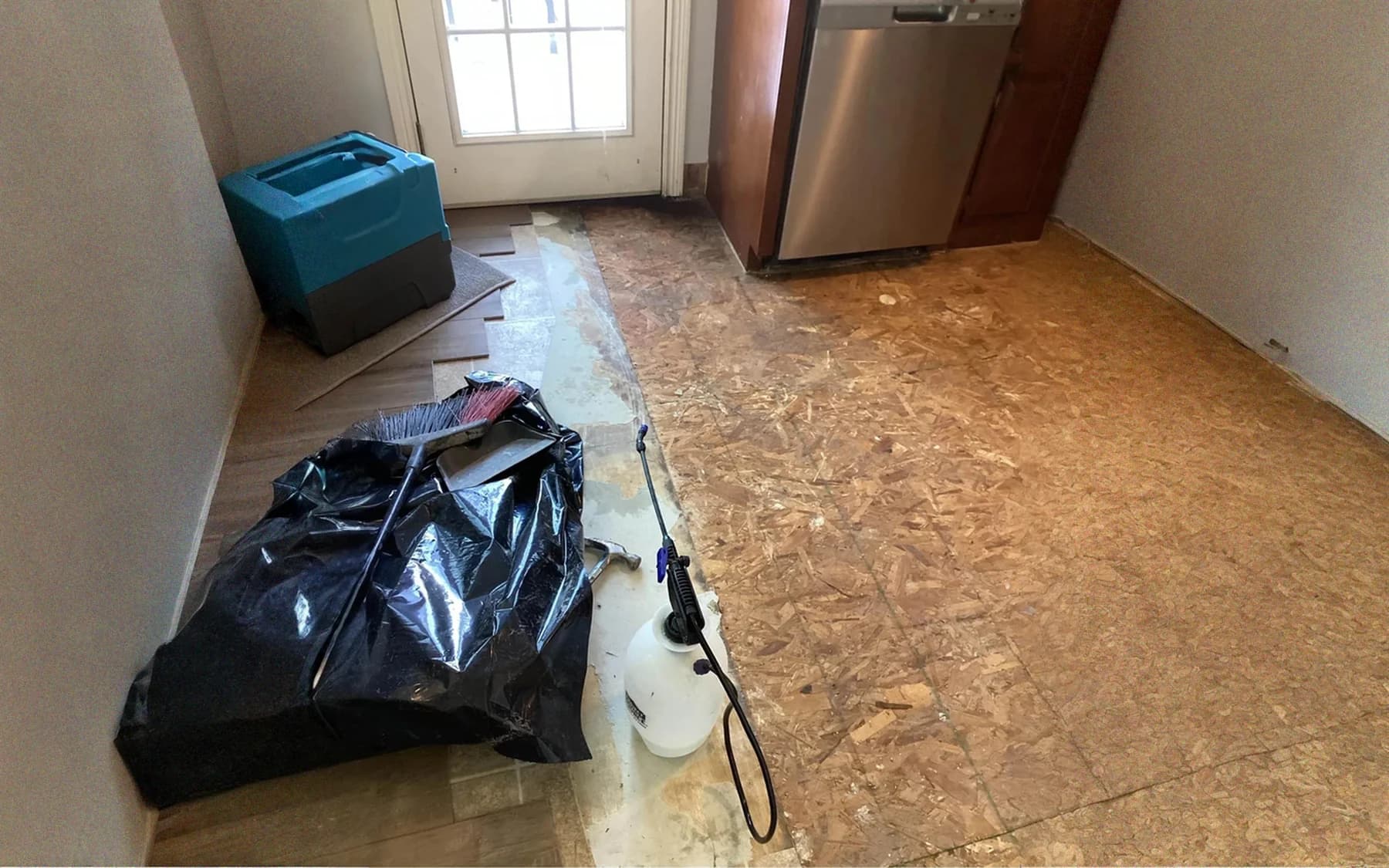A pump sprayer standing on exposed subfloor beside a waste bag, ready to apply antimicrobial treatment after the flooring came out